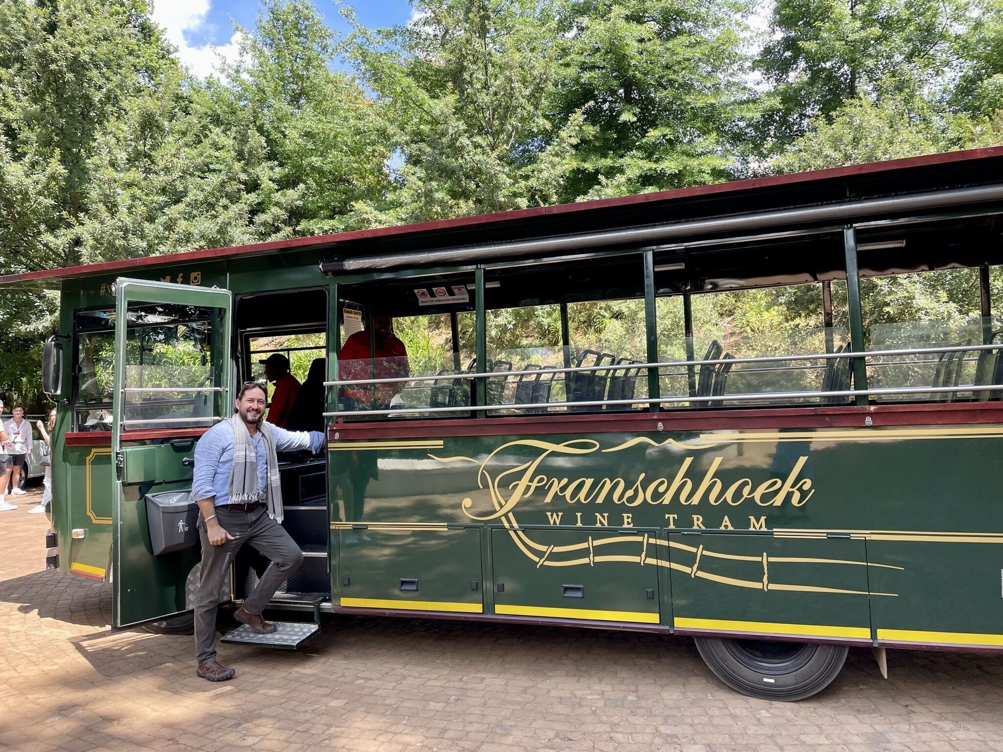 Ein grüner Straßenbahnwagen mit der Aufschrift "Franschhoek Wine Tram", geparkt im Freien, umgeben von Personen.