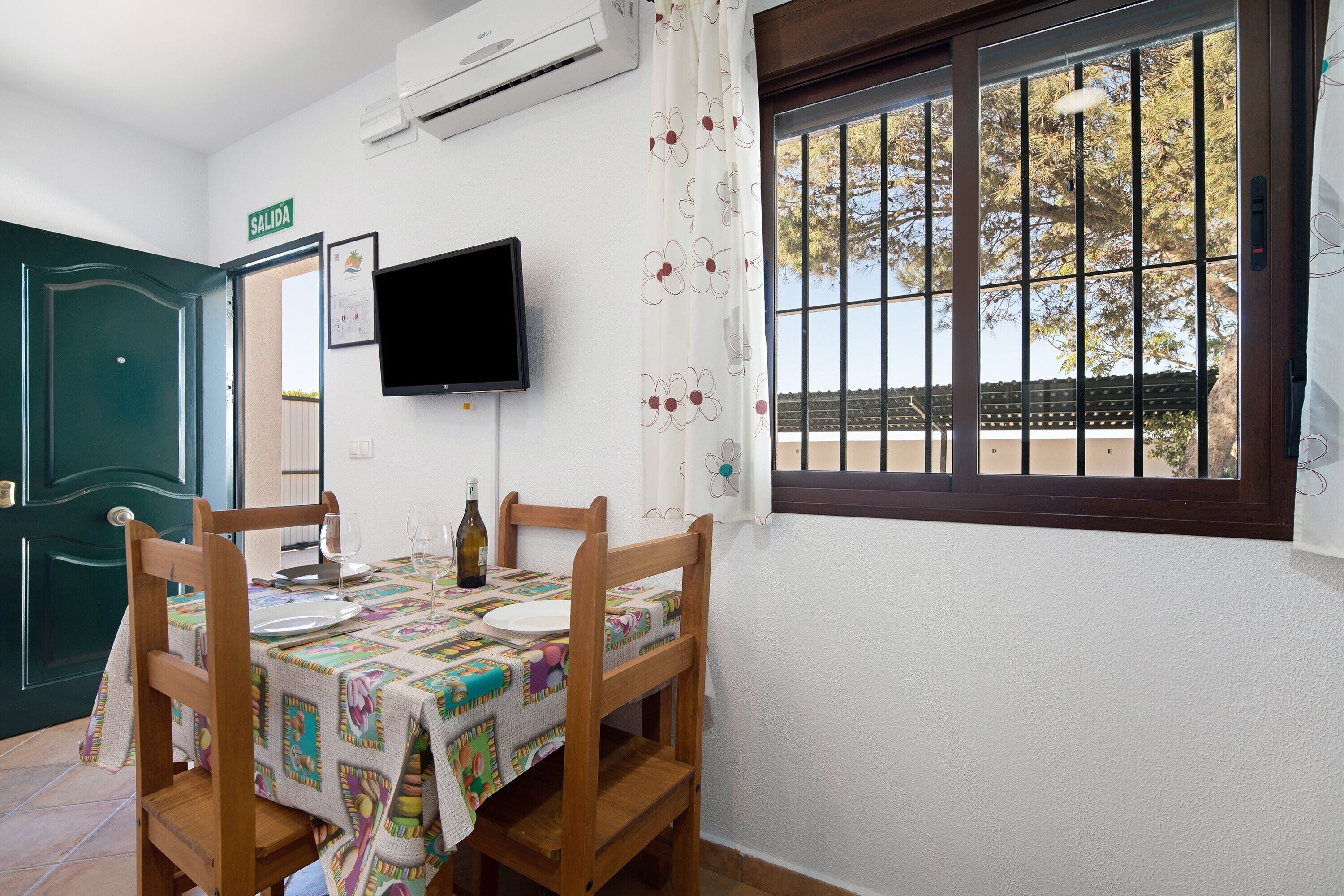 A dining area with a table set for four, a television mounted on the wall, and a window with bars.