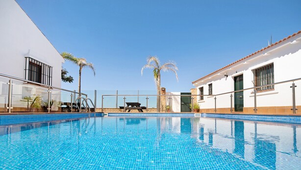 A swimming pool with clear blue water, surrounded by a white building with a tiled roof, palm trees, and a glass railing. - Holiday Home "Novobarrosa A" with Sea View, Shared Pool & Wi-Fi (Chiclana de la Frontera)
