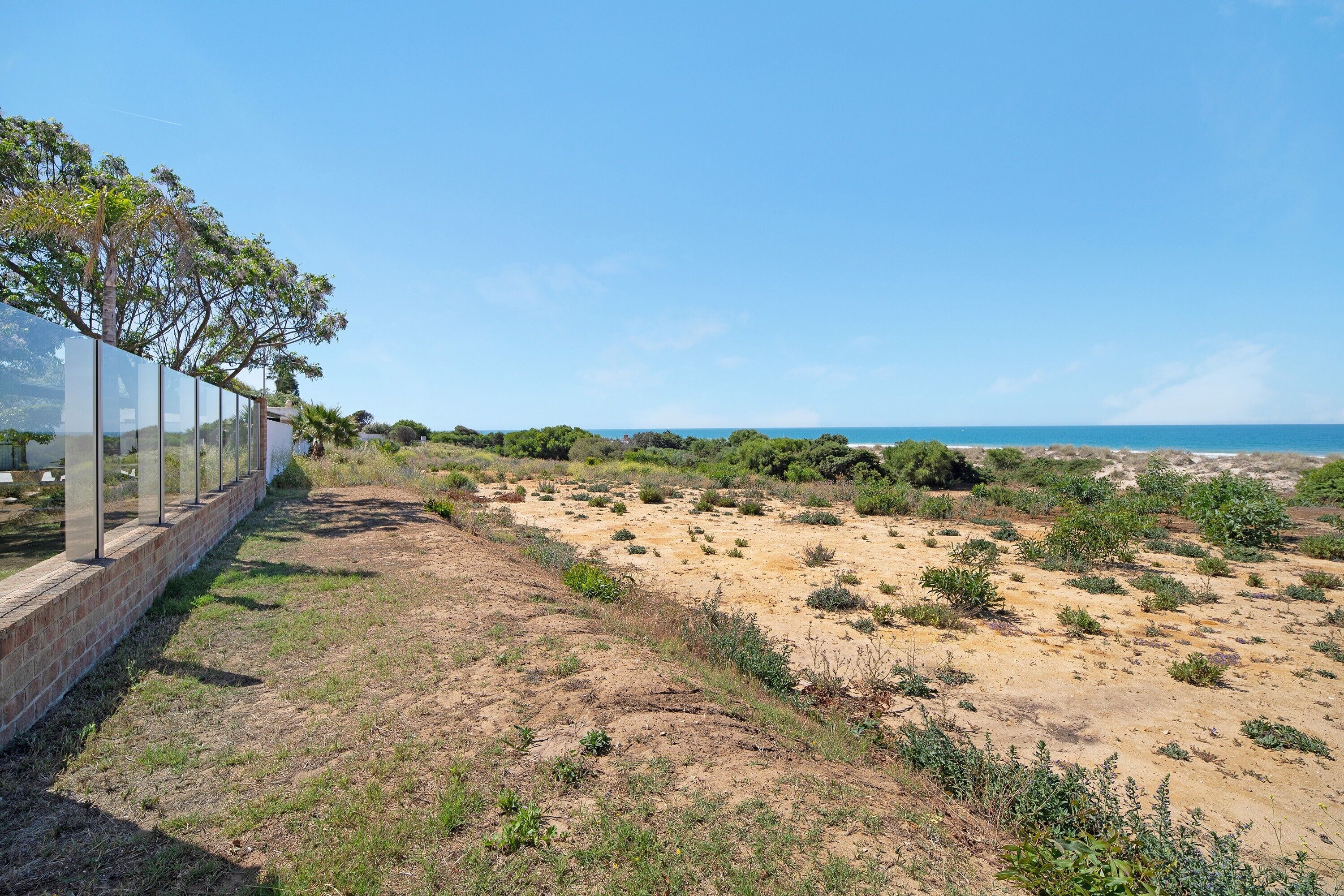 Eine Küstenlandschaft mit blauem Himmel, einem Sandstrand mit vereinzelter Vegetation und einer Struktur mit Glasbarriere auf der linken Seite.