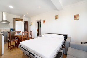 A bedroom with a bed, bedside table, and a chair. There is a kitchenette with a stove and a refrigerator.