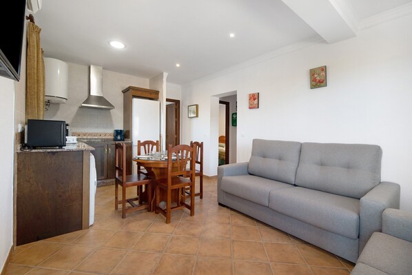A living room with a grey sofa, a dining table with chairs, a kitchen area with a stove, and a TV.