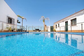Una piscina con agua azul cristalina, rodeada por un edificio blanco con tejado de baldosas, palmeras y una barandilla de vidrio.