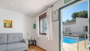 A room with a grey sofa, a small table, a window with curtains, and a view of an outdoor swimming pool.