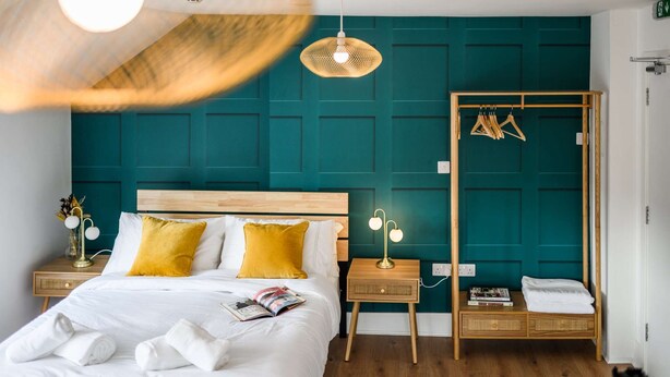 A neatly made bed with white and yellow pillows, a bedside table with a lamp, and a wooden wardrobe with clothes hanging inside.