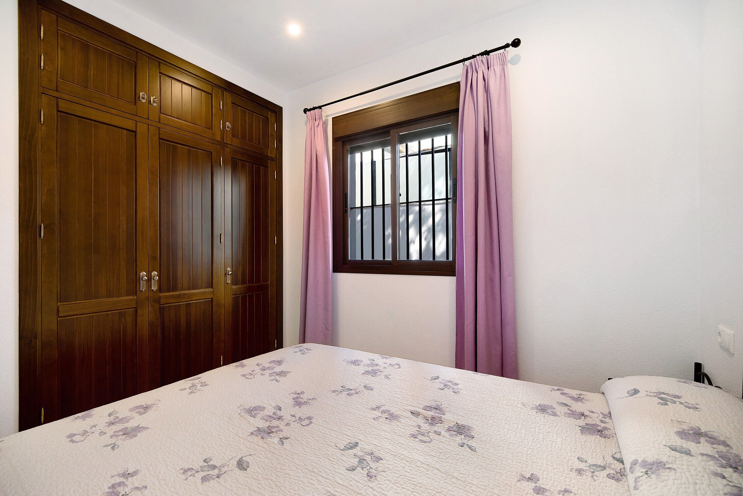Un dormitorio con cama, armario de madera, ventana con barrotes y cortinas rosadas.