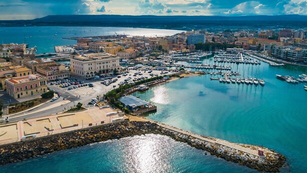 Vista aérea de una ciudad costera con un puerto deportivo, edificios y un litoral rocoso.