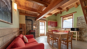 A cozy interior with a wooden ceiling, a dining area with a table and chairs, a red sofa, and a kitchen with red cabinets and a stone wall. - Chalet 'Civico 2' with Mountain View, Balcony and Wi-Fi (Marmora)