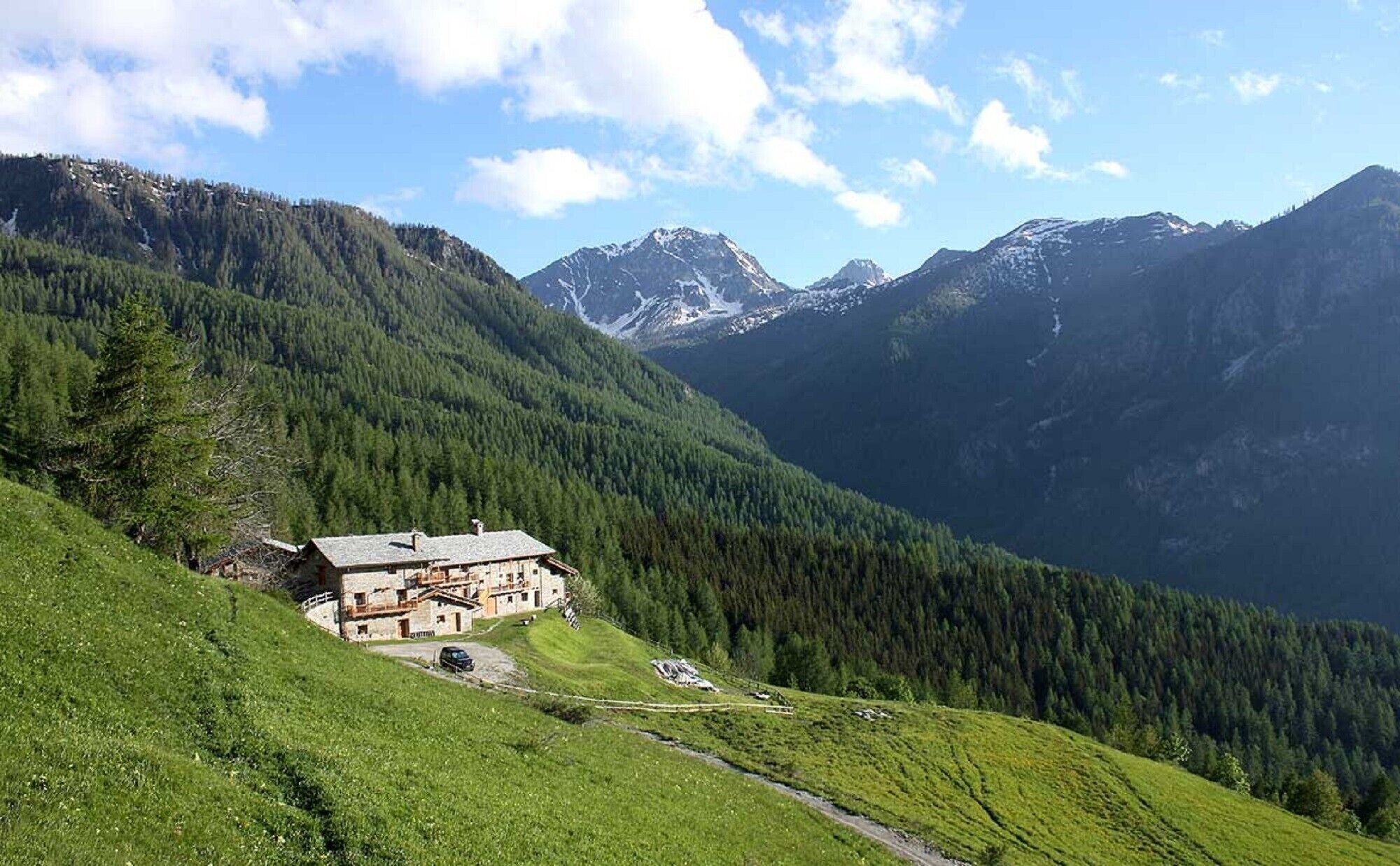 A mountain lodge with a stone facade, surrounded by lush greenery and snow-capped peaks.