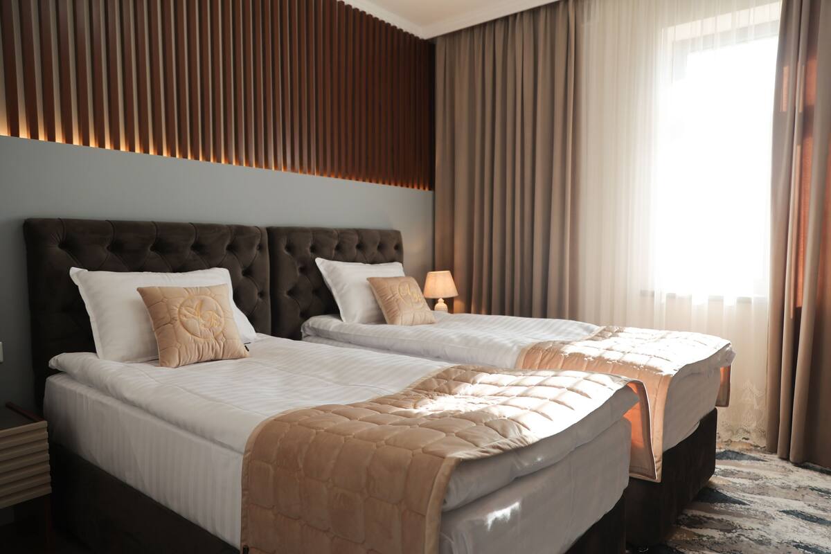 A hotel room with two beds, a large window with curtains, a wall-mounted panel, and a bedside lamp.