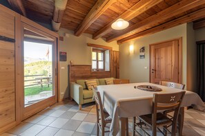 A wooden interior with a dining table, chairs, a sofa, and a window with a view of a wooden deck.