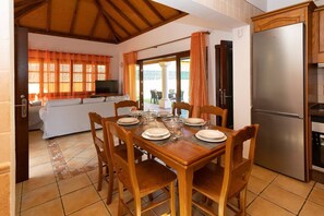 A dining area with a wooden table set for four, a sofa, a TV, and a kitchen with a refrigerator.