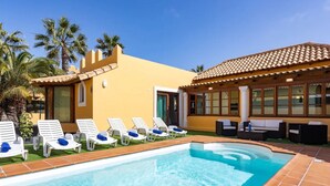 Un espace piscine avec des chaises longues, un petit bâtiment doté d’un toit en tuiles et des palmiers.