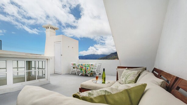 A bedroom with a bed, pillows, a table with a bottle and glass, and a view of mountains.