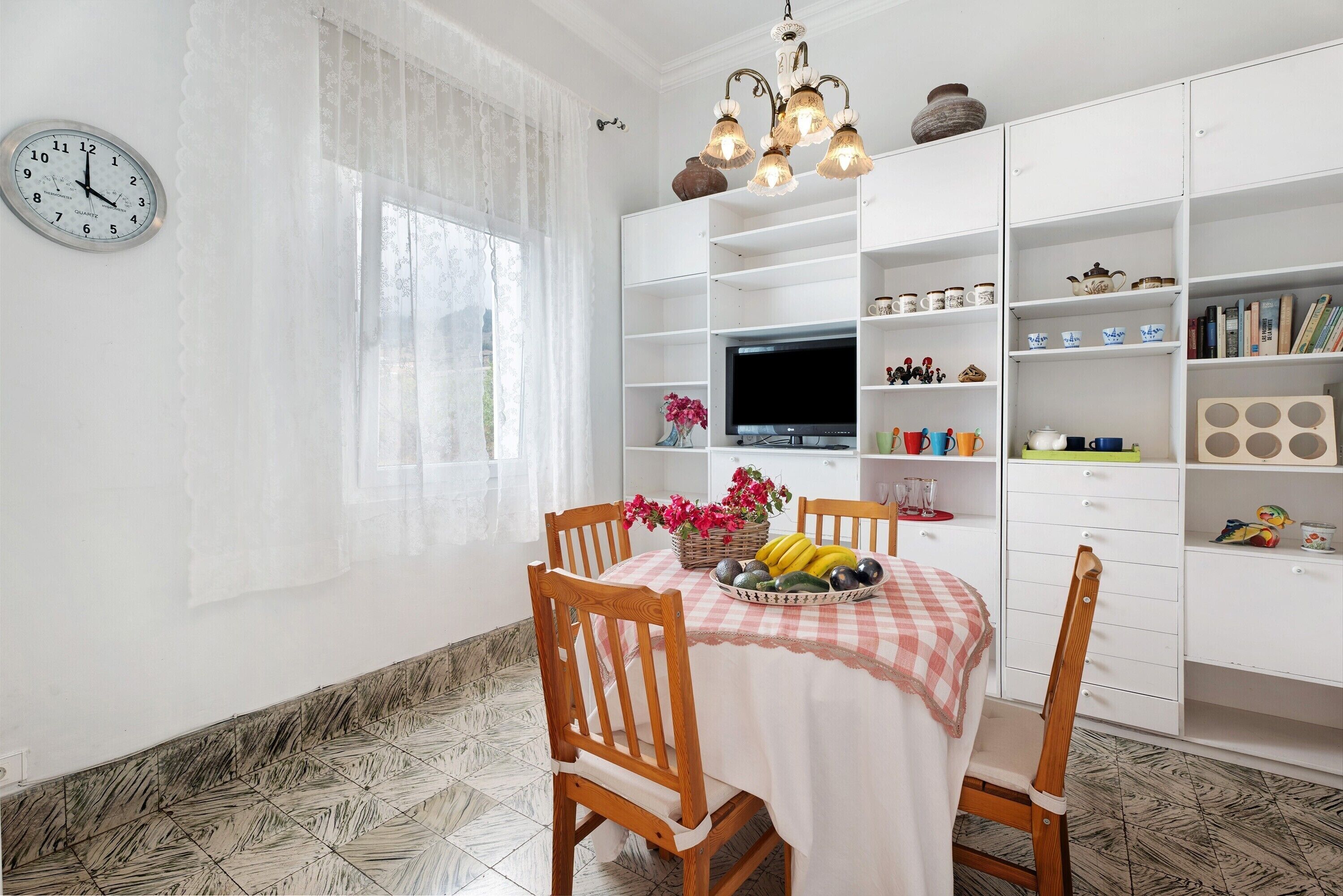 Un comedor con una mesa dispuesta para dos, un televisor sobre un mueble bajo y un armario blanco con estantes.