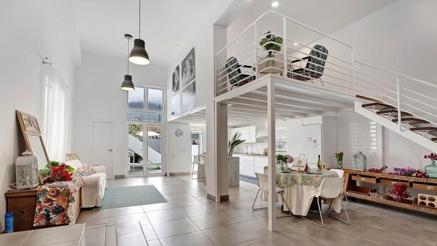 A spacious living area with a high ceiling, a dining table set for four, a sofa, and a staircase leading to an upper level.