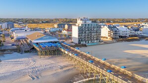 A coastal hotel with a wooden pier extending into the water, a beach area with umbrellas, and a view of the ocean.