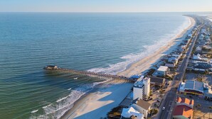 An aerial view of a coastal town with a long pier extending into the sea, adjacent to a sandy beach and lined with buildings.