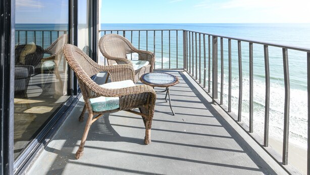 A balcony with wicker furniture and a small round table, overlooking a beach with a clear view of the ocean.
