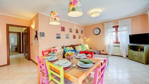 Un coin repas avec une table en bois dressée pour quatre personnes, des chaises colorées, un canapé, un téléviseur et une horloge au mur.