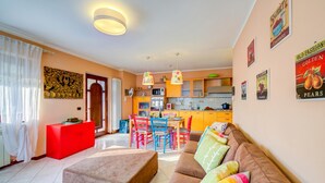 Un salon moderne avec un canapé, un pouf et des coussins colorés. On aperçoit, au fond, une cuisine avec un coin repas.