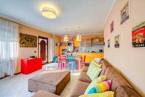 A modern living room with a sofa, ottoman, and colorful cushions. A kitchen with a dining area is visible in the background. - Casa Angiolina Lake & Mountain View (Tremosine)