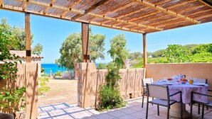 Un patio techado al aire libre con una mesa lista para comer, rodeado de sillas, y vistas panorámicas del mar y la vegetación.