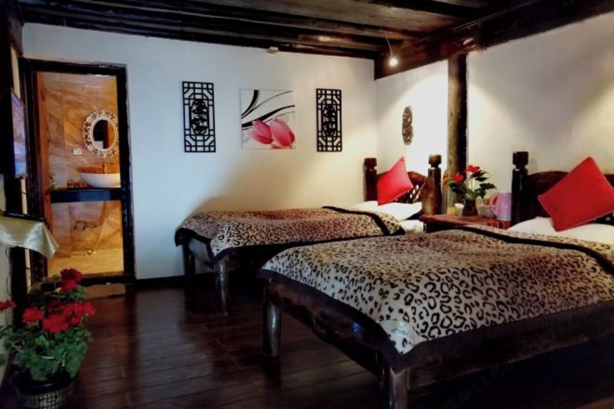 A room with two beds, each with a leopard print bedspread and a red pillow. There is a wooden door with a mirror and a small table with a vase of flowers. The walls are adorned with framed artwork.