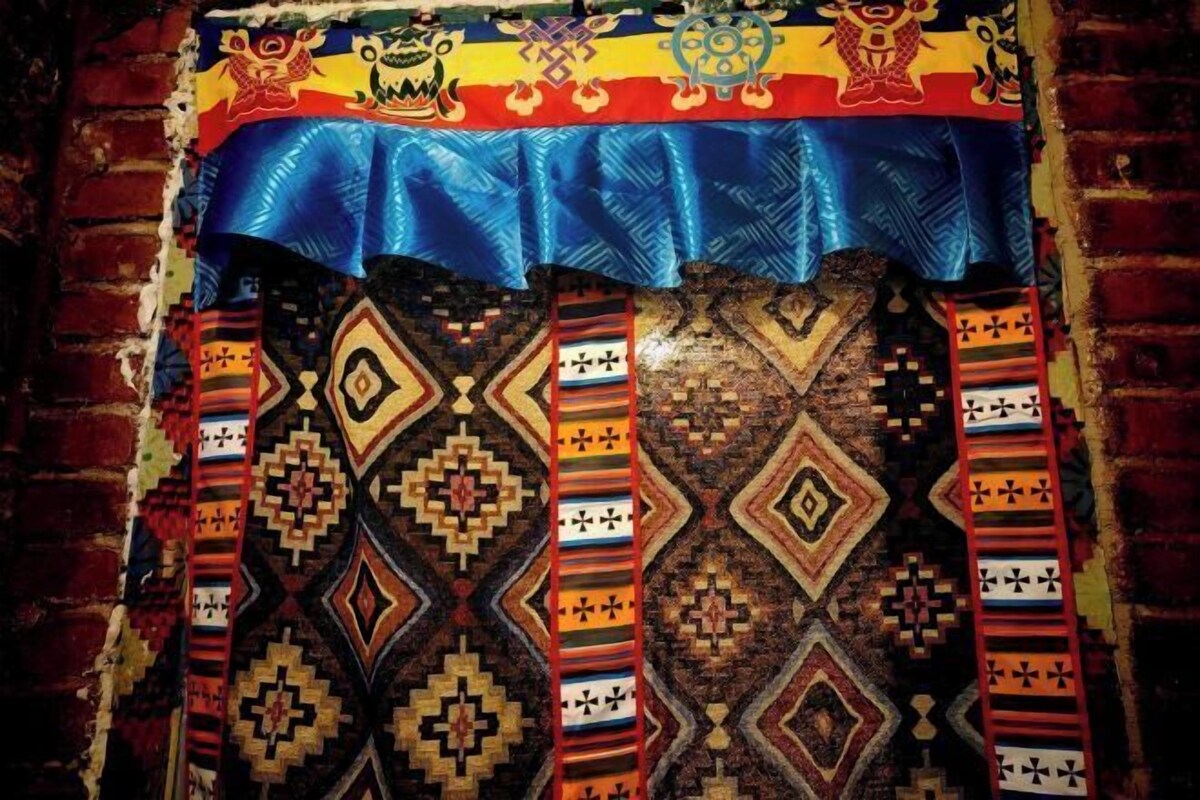 A traditional woven textile with geometric patterns and a blue border, displayed against a brick wall.