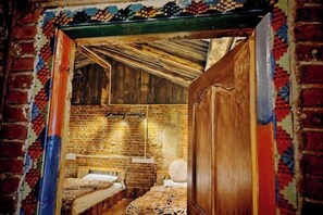 A rustic room with exposed brick walls, wooden beams, and a traditional door. There are two beds with patterned bedding.