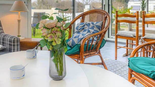 Uma sala de estar moderna com uma mesa redonda, cadeiras de vime, um vaso com flores e uma vista através de grandes janelas.