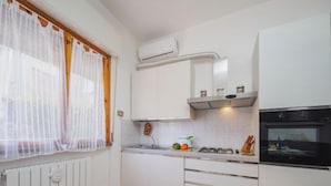 Une cuisine avec des meubles blancs, une cuisinière, un four, une fenêtre avec des rideaux légers et un radiateur.