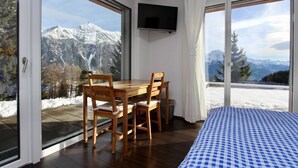 Una habitación con una mesa y sillas de madera, una cama con colcha a cuadros, televisor montado y ventanas amplias con vistas a la montaña.