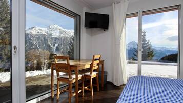Une pièce avec une table et des chaises en bois, un lit recouvert d’une courtepointe à carreaux, un téléviseur fixé au mur et de grandes fenêtres offrant une vue sur la montagne.