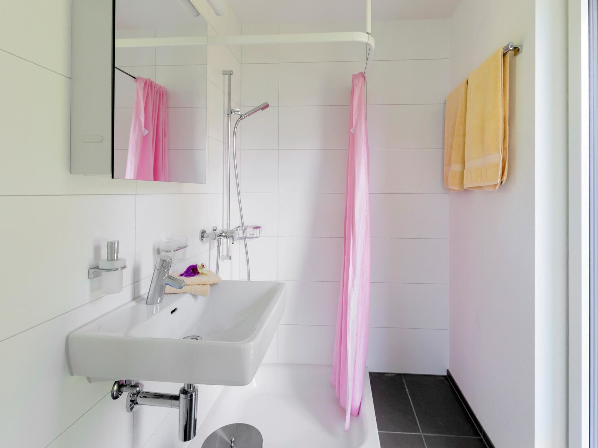 Une salle de bain moderne avec un lavabo blanc, un miroir, une douche avec un rideau rose et une serviette jaune accrochée au mur.