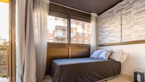 Une chambre à coucher avec un lit, des rideaux, une fenêtre donnant sur des immeubles et un mur sur lequel sont inscrits des noms de villes.