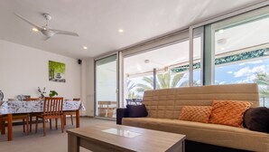 A modern living room with a sofa, dining table, and chairs. There is a ceiling fan, a large window with a view of palm trees, and a painting on the wall. - Acantilados by Interhome (Salou)
