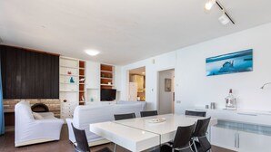 A modern kitchen with white cabinetry, a dining area with a white table and black chairs, and a living area with a white sofa and a TV. - Acantilados by Interhome (Salou)