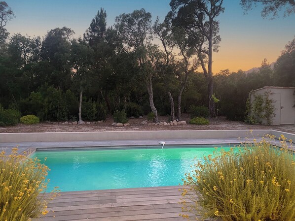 Una piscina con plataforma de madera, rodeada de árboles y un cobertizo pequeño.