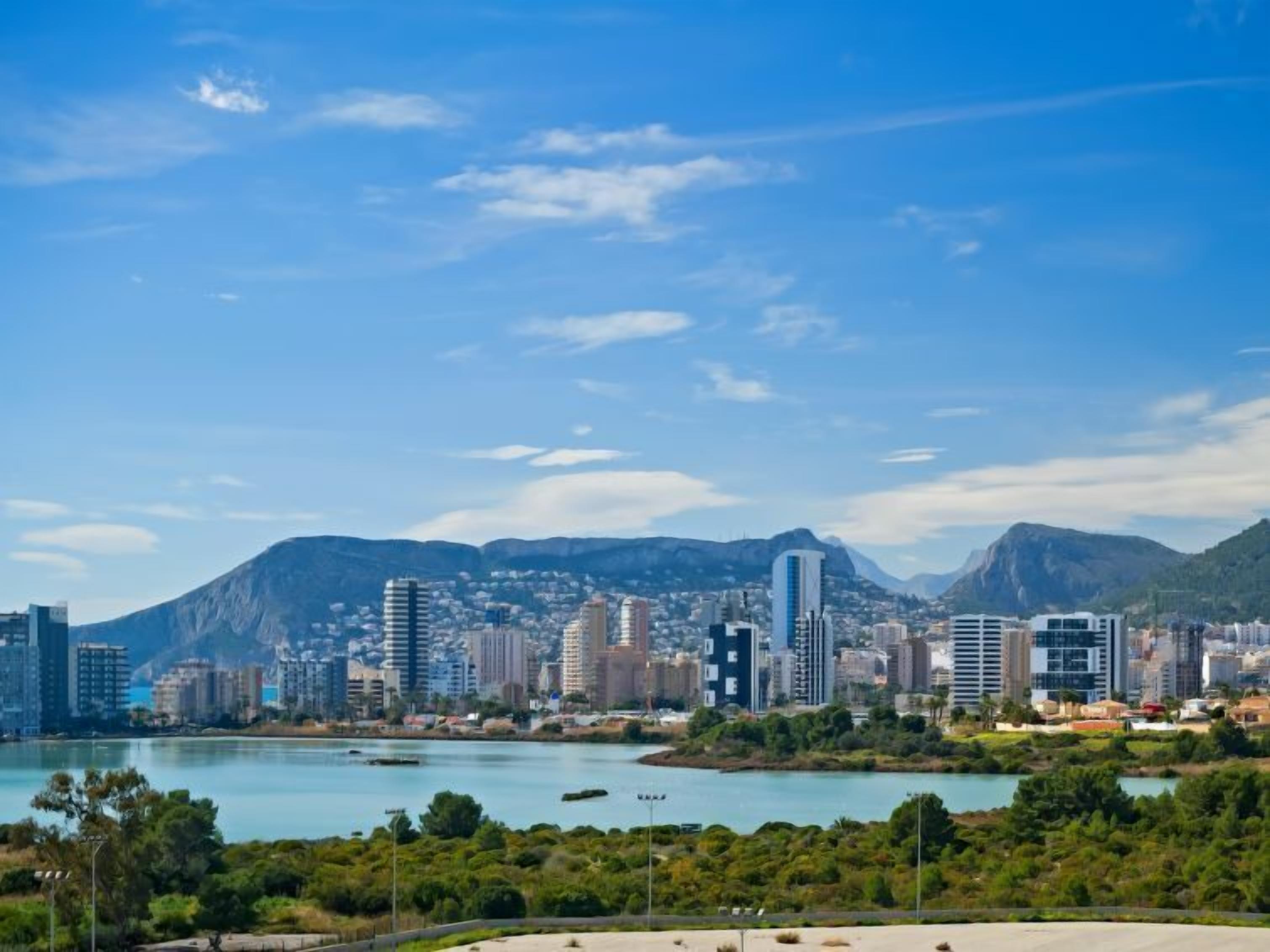 A coastal cityscape with modern buildings, a clear blue sky, and a mountain in the background.
