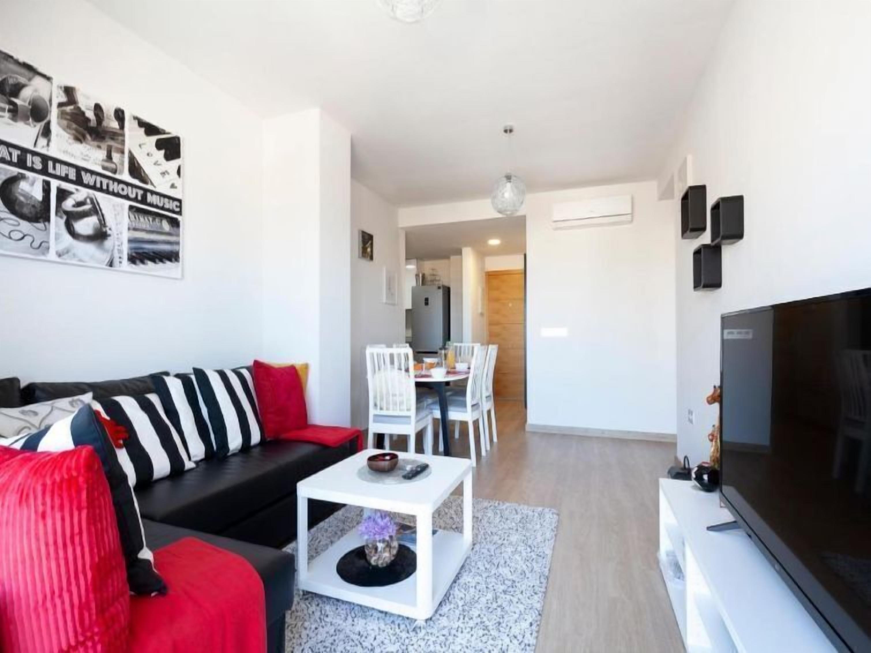 Een moderne woonkamer met een zwarte bank, rode kussens, een witte salontafel en een flatscreen televisie.
