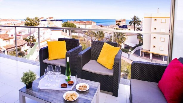 Een balkon met rieten meubilair, een tafel met glazen en een fles, en uitzicht op zee en gebouwen.