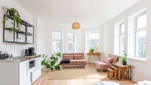Ein moderner, gut beleuchteter Wohnraum mit Küchenzeile, einer Couch, einem Essbereich und großen Fenstern, die viel Tageslicht hereinlassen.