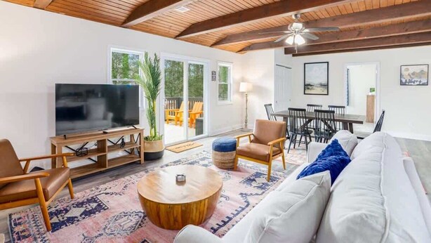Una sala de estar amplia con un televisor grande, vigas de madera en el techo, un sofá y un comedor con sillas y una mesa.