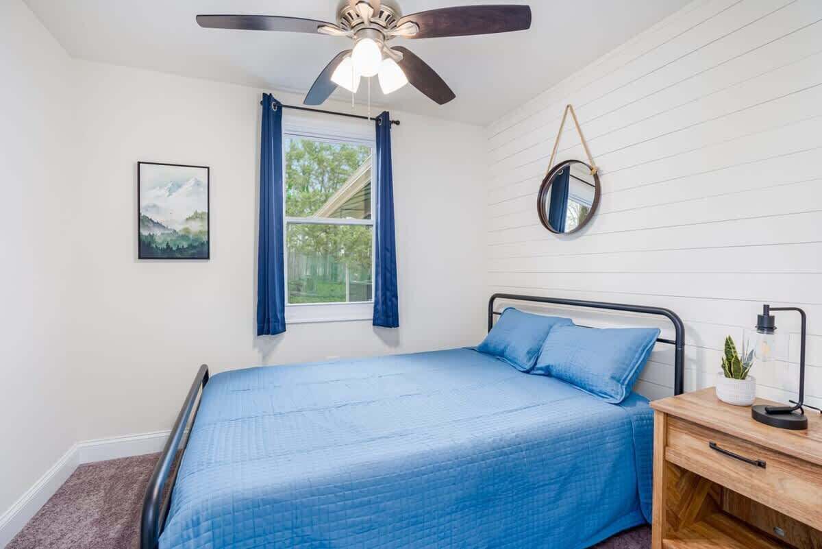 A bedroom with a bed, a ceiling fan, a window with curtains, a bedside table with a lamp, and a wall-mounted mirror.