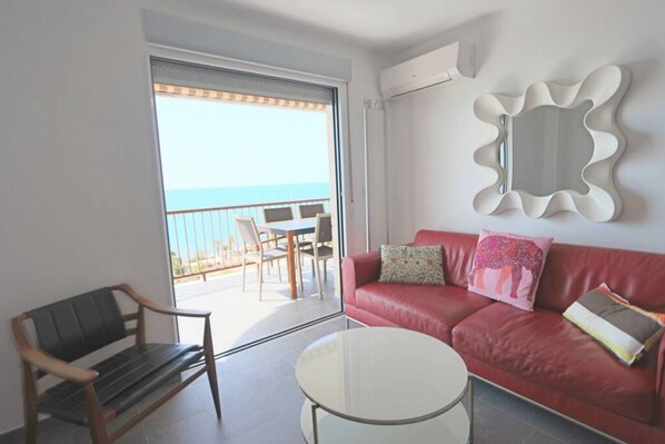 Una sala de estar moderna con un sofá rojo, una mesa de centro de vidrio y un banco. Hay un balcón con vistas al mar y una mesa con sillas afuera.