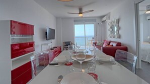 Un espace repas moderne avec une table blanche dressée pour un repas, des meubles de couleur rouge et une vue sur la mer grâce à de grandes fenêtres.