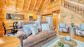 L’intérieur d’un chalet confortable, avec des murs en bois, un canapé, des fauteuils, une table à manger et un escalier.
