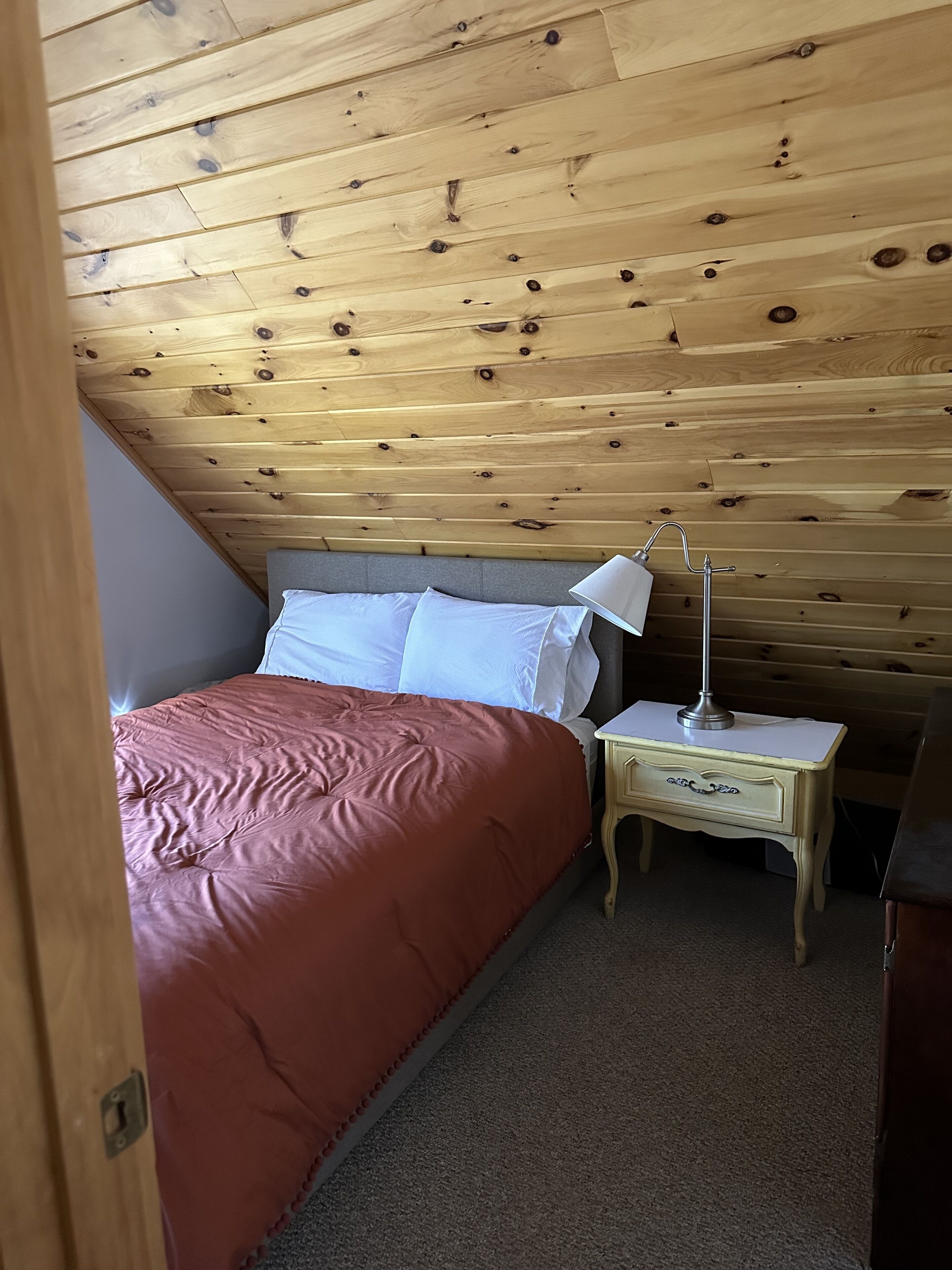A bedroom with a wooden ceiling, a bed with a red blanket, white pillows, and a bedside table with a lamp.