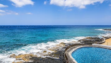 A coastal pool area with clear blue water, surrounded by rocky terrain and a view of the ocean.